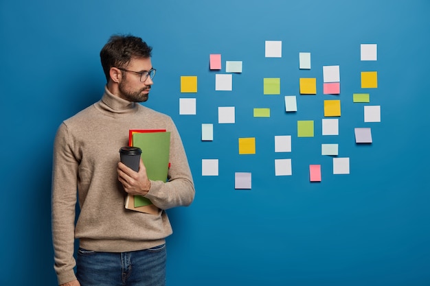 Foto grátis freelancer ou estudante masculino lê ideias escritas em notas de papel coladas na parede azul, segura café para viagem e um bloco de notas, aprende palavras estrangeiras com adesivos coloridos