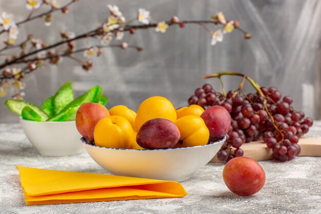 Frente, damascos doces, frutas amarelas dentro do prato com ameixas e uvas na mesa branca