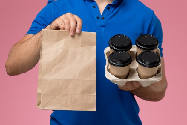 Frente de perto, mensageiro masculino de uniforme azul segurando pacotes de comida em copos de café de entrega na rosa, entrega de trabalho de serviço uniforme