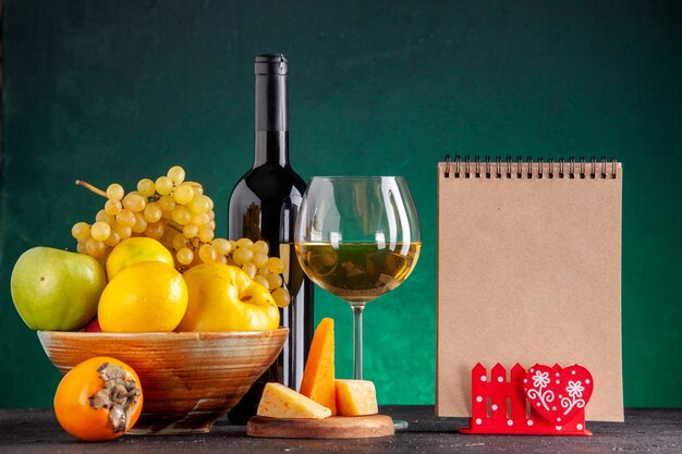 Frente frutas frescas em uma tigela de madeira maçãs marmelo uvas limão garrafa de vinho de caqui e queijo de vidro no bloco de notas de placa de madeira na mesa verde