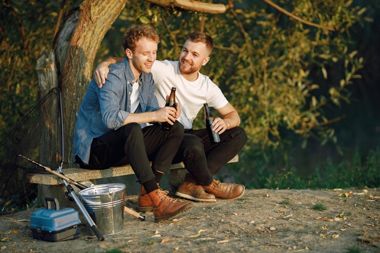 Foto grátis friends está sentado perto de uma árvore e pescando. dois pescadores conversando e bebendo cerveja
