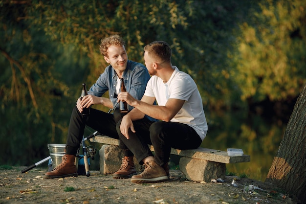 Friends está sentado perto de uma árvore e pescando. Dois pescadores conversando e bebendo cerveja