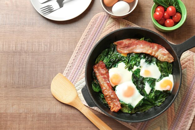 Frigideira com ovos saborosos, espinafre e bacon na mesa de madeira