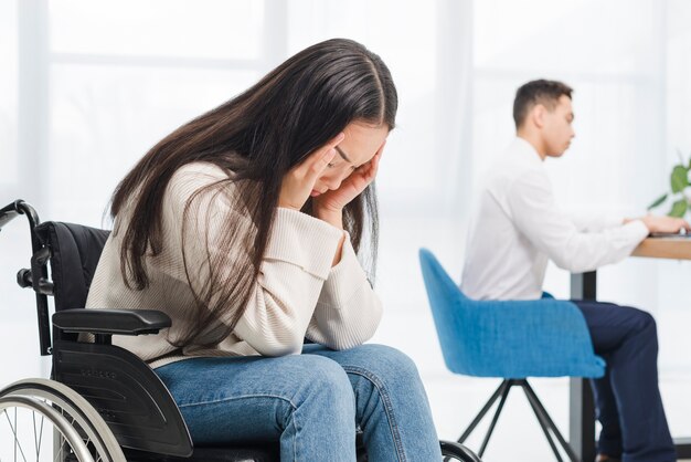 Frustrado, mulher jovem, sofrimento, de, dor de cabeça, sentando, ligado, cadeira roda, frente, homem negócios, trabalhando, em, escritório