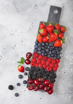 Frutas orgânicas frescas de verão se misturam na placa de mármore preta sobre fundo claro de mesa de cozinha. framboesas, morangos, mirtilos, amoras e cerejas. vista do topo