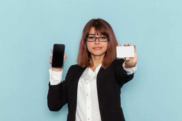 Funcionária de escritório feminina de terno estrito segurando o telefone e um cartão branco na superfície azul.