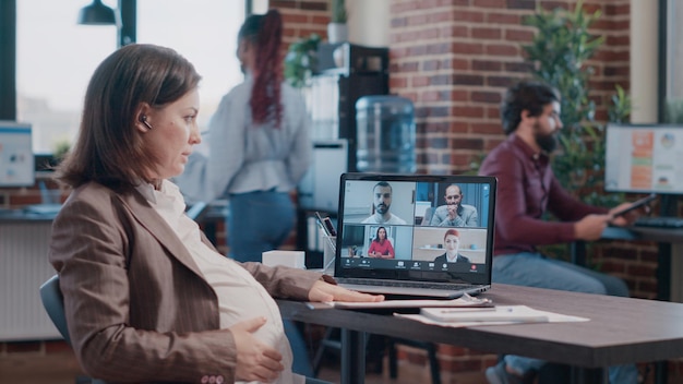 Funcionária grávida usando videochamada comercial no laptop para conversar com colegas de trabalho. Mulher esperando bebê e participando de reunião em videoconferência remota online, falando sobre planejamento de projetos.