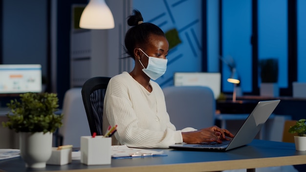 Funcionário africano com máscara facial de proteção trabalhando horas extras com laptop no novo escritório normal de negócios para respeitar o prazo do projeto, analisando documentos sentados na mesa durante horas extras durante a pandemia global