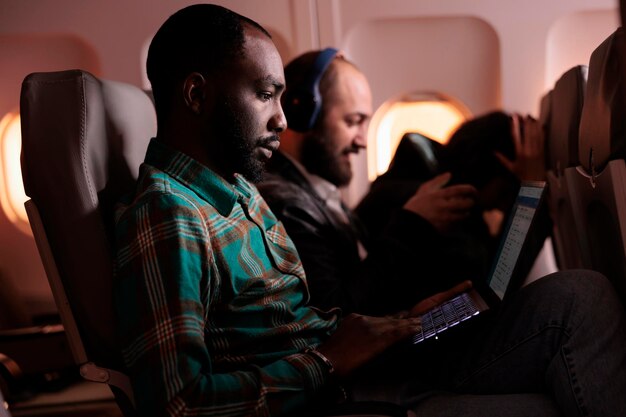 Funcionário do sexo masculino viajando em classe econômica e usando laptop durante o voo, voando para o exterior em destino de férias ou viagem de trabalho. Trabalhando no computador durante o pôr do sol, transporte aéreo.