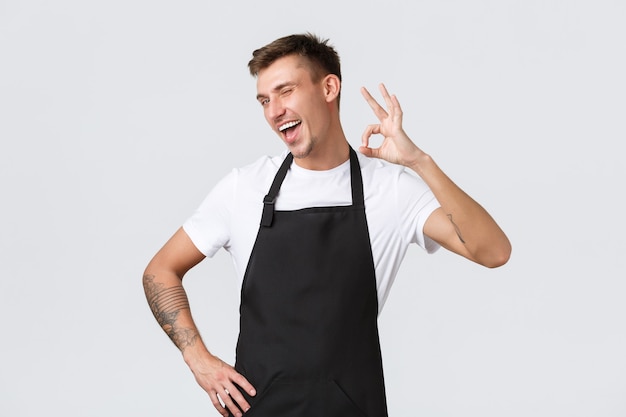 Foto grátis funcionários, mercearias e conceito de cafeteria. barista extrovertido e alegre, garçom de avental preto garante qualidade, gesto de aprovação e piscadela atrevida, aprovar e recomendar café