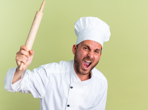 Furioso jovem cozinheiro caucasiano com uniforme de chef e boné gritando segurando o rolo de massa