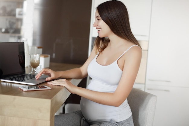 Futura mãe sendo feliz e alegre verificando a caixa de correio via laptop retrato de mulher grávida despreocupada sentada na cozinha perto de notebook e smartphone escrevendo blog sobre gravidez para mulheres copiar espaço