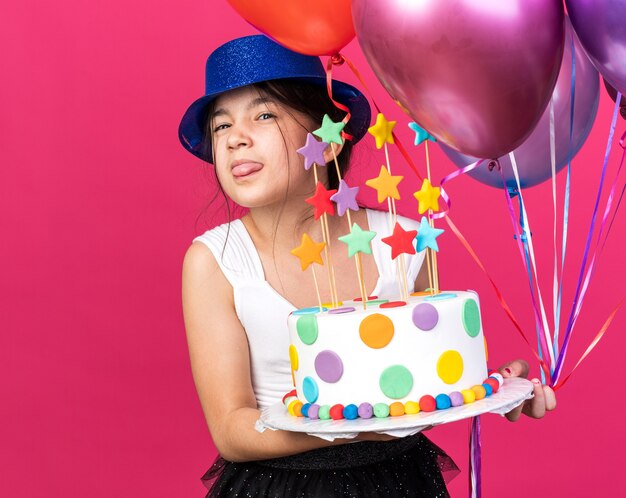 gananciosa jovem caucasiana com chapéu de festa azul estica a língua segurando um bolo de aniversário e balões de hélio isolados na parede rosa com espaço de cópia
