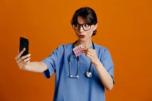 Ganancioso toma uma selfie segurando pílulas jovem médica vestindo uniforme quinto estetoscópio isolado em fundo laranja