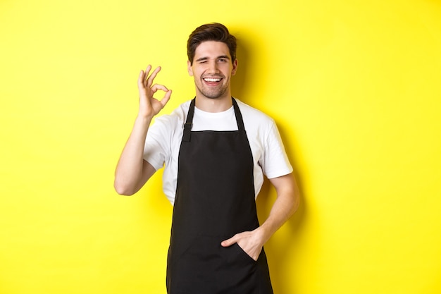 Garçom confiante e bonito mostrando sinal de ok usando avental preto e de pé contra o fundo amarelo ...