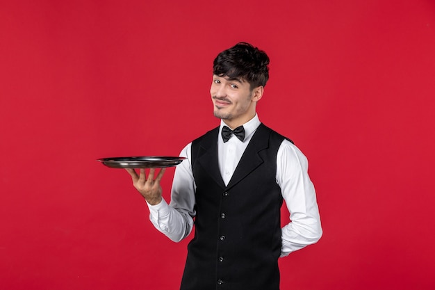 Garçom jovem confiante e sorridente com um uniforme amarrando uma borboleta no pescoço segurando a bandeja no fundo vermelho isolado