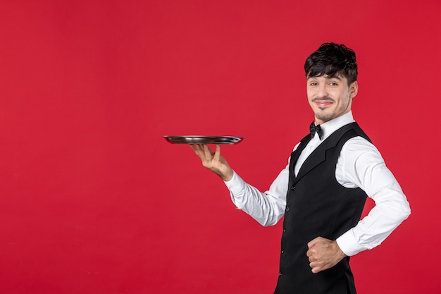 Garçom jovem orgulhoso e confiante em um uniforme amarrando uma borboleta no pescoço segurando uma bandeja no fundo vermelho isolado