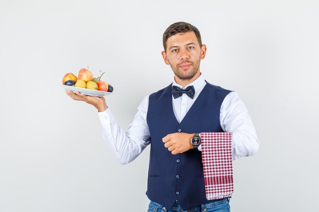 Garçom segurando o prato cheio de frutas em camisa, colete, jeans, vista frontal.