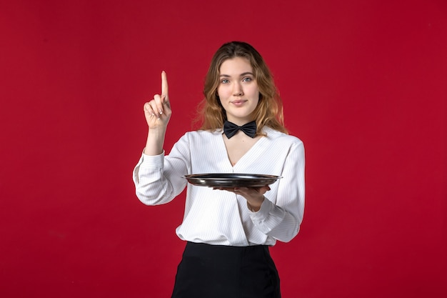 Foto grátis garçonete confiante mulher borboleta no pescoço segurando a bandeja apontando para cima no fundo vermelho