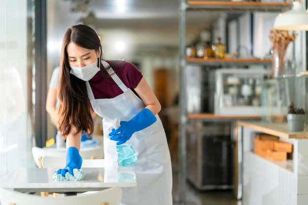 Garçonete de equipe asiática usando avental de máscara facial de proteção e mesa de limpeza de luva de borracha com spray de álcool desinfetante para prevenir infecção coronavírus no café café novo conceito normal