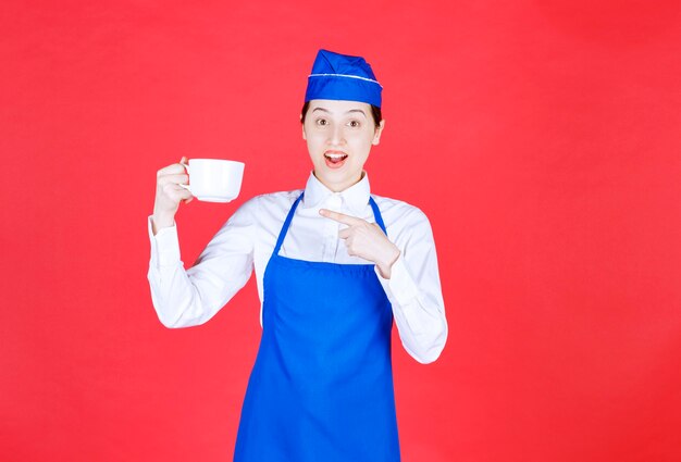 Garçonete de mulher de uniforme em pé e apontando para uma xícara na parede vermelha.