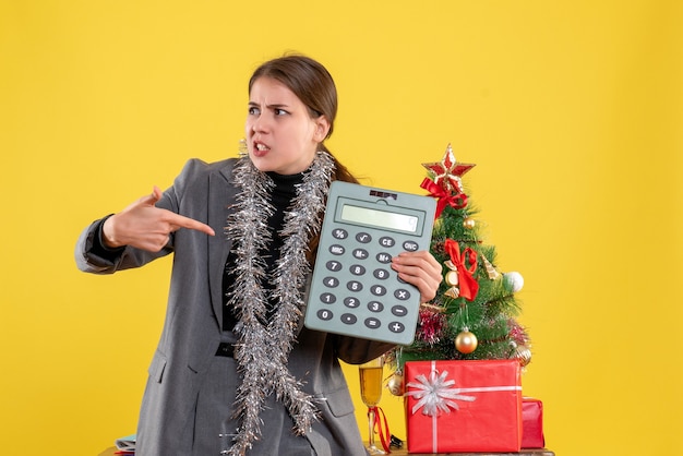 Garota admirada de frente mostrando a calculadora perto da árvore de natal e um coquetel de presentes
