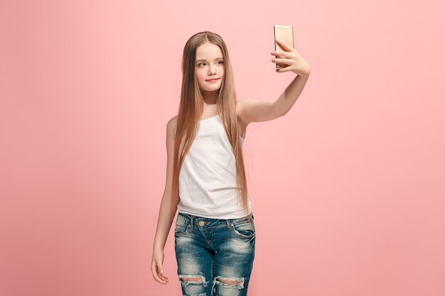 Garota adolescente feliz em pé, sorrindo na parede rosa, tirando foto de selfie pelo telefone celular. Emoções humanas, conceito de expressão facial. Vista frontal.