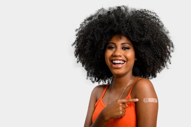Garota adolescente negra sorri e aponta para o braço com adesivo de vacina, ela não usa protetor facial, isolado em fundo cinza.