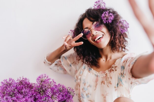 Garota africana relaxada tirando foto de si mesma com flores roxas. Foto interna do fascinante modelo feminino encaracolado em óculos de sol.