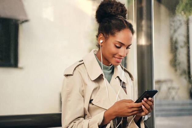 Garota afro-americana casual muito sorridente em elegante casaco de trincheira e fones de ouvido alegremente usando celular no ponto de ônibus