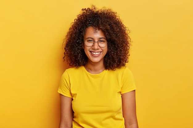 Garota afro-americana divertida e alegre ri durante uma conversa divertida, sorri amplamente, mostra os dentes brancos, usa óculos transparentes e camiseta amarela, sorri com os dentes, encoraja a expressão