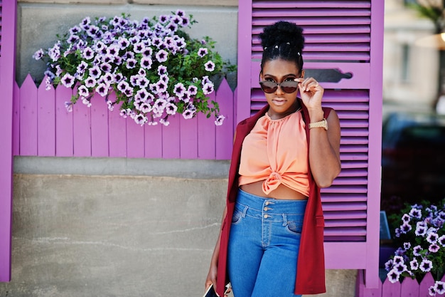 Garota afro-americana em óculos de sol posou contra janelas roxas com telefone celular à mão