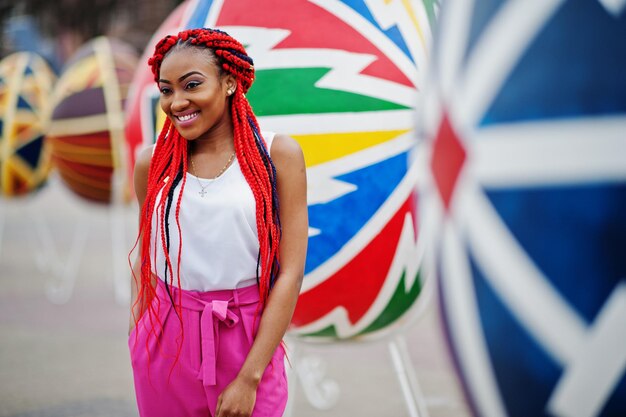 Garota afro-americana na moda em calças cor de rosa e dreads vermelhos posou ao ar livre contra ovos grandes