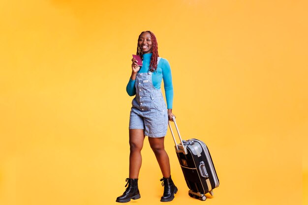 Garota afro-americana segurando passe de identificação antes de embarcar em voo de avião, viagem de férias no exterior. Mulher alegre partindo em fuga internacional, destino com malas e passaporte.