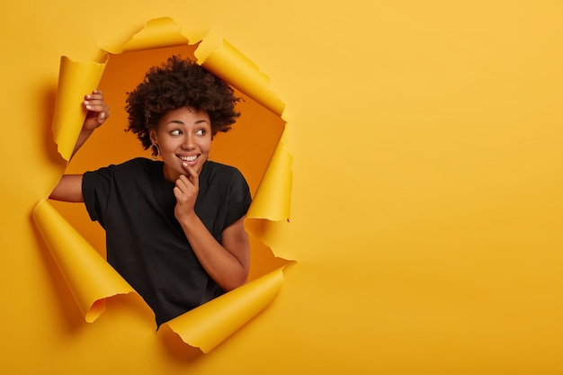 Foto grátis garota afro positiva segura o queixo, focada de lado, se sente cativante e alegre, usa uma camiseta casual, posa sozinha no buraco do papel