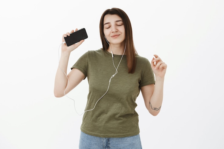 Foto grátis garota alcançando o nirvana e emoções positivas, tendo vibrações positivas com o som incrível de fones de ouvido, ouvindo música dançando sensualmente com os olhos fechados e um sorriso feliz, levantando a mão com o smartphone
