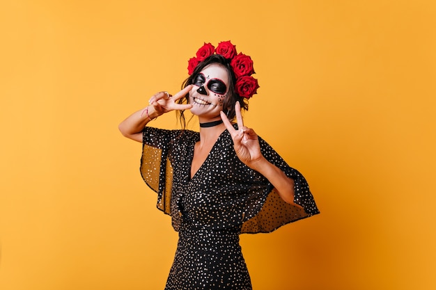 Foto grátis garota alegre e animada para o halloween está posando na parede laranja, mostrando o símbolo da paz. retrato de mulher de vestido preto fofo sorrindo