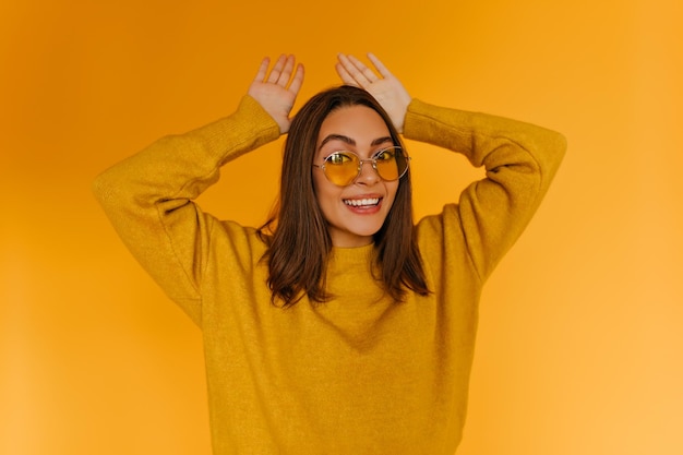 Garota alegre positiva em óculos de sol amarelos se divertindo durante a sessão de fotos Linda senhora ativa no suéter relaxando no estúdio