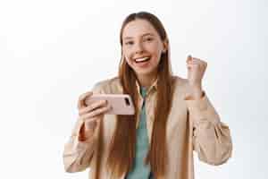 Foto grátis garota alegre se regozijando comemorando conquistas no aplicativo móvel segurando o smartphone horizontal e fazendo a bomba de punho sorrindo satisfeita em alcançar a meta no fundo branco do aplicativo
