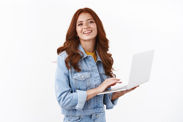 Foto grátis garota ambiciosa e bonita lidera vanlife trabalhando remoto em pé sobre fundo branco elegante jaqueta jeans segure laptop sorrindo satisfeito freelancing escrevendo ensaio universitário