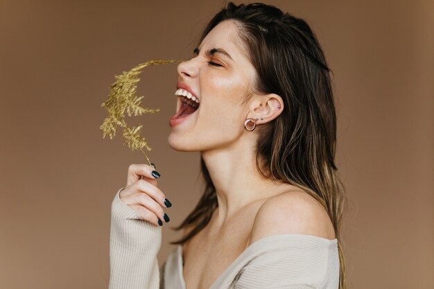 Garota animada em brincos posando com flor. Jovem morena inspirada rindo durante as filmagens.