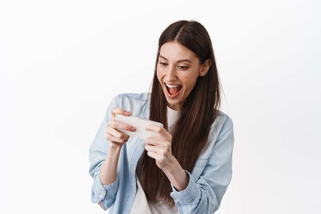 Garota animada jogando videogame de corrida no smartphone, jogando no celular, olhando para a tela com alegria e sorrindo, fique de pé sobre um fundo branco