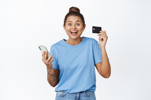 Garota animada segurando cartão de crédito e celular, pagando com aplicativo bancário, compras online no smartphone, em pé contra um fundo branco