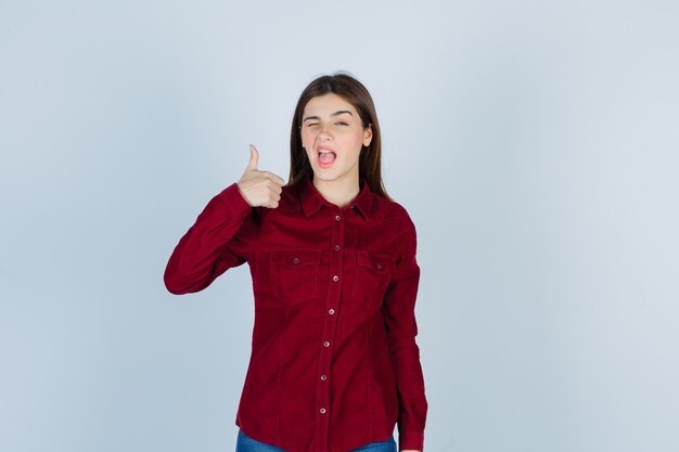 garota aparecendo o polegar enquanto pisca em uma camisa cor de vinho e parece satisfeita.