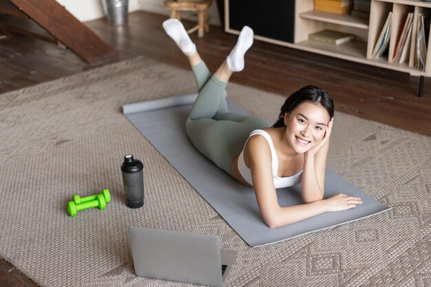 Garota asiática fitness tendo a pausa do treino, exercitando-se em casa no tapete com halteres e prote ...