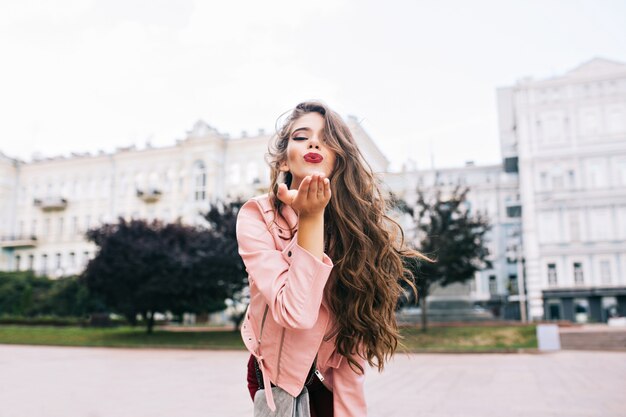 Garota atraente com penteado longo, se divertindo na cidade. Ela tem uma jaqueta rosa, mandando um beijo com lábios carnudos.