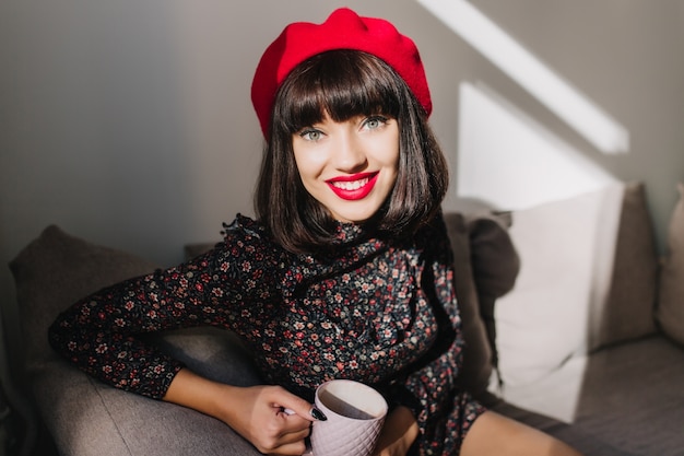 Garota atraente de cabelos escuros na boina vermelha e vestido retrô, sentada numa sala aconchegante enquanto bebia chá. Retrato de uma adorável jovem morena com roupa francesa, posando com prazer durante a pausa para o café em casa