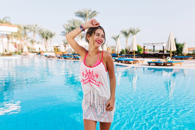 Garota atraente e bem torneada usando minissaia e camiseta regata de franjas dançando com a mão para cima esperando a diversão na piscina
