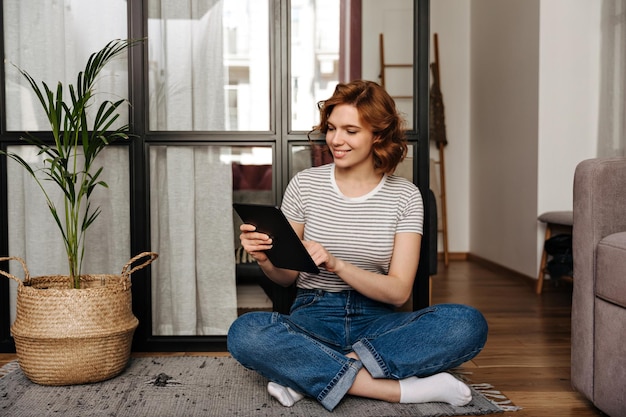 Foto grátis garota atraente em camiseta listrada com sorriso funciona em tablet enquanto está sentado na sala de estar