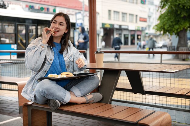 Garota atraente em estilo casual come um hambúrguer com café sentada na esplanada de um café